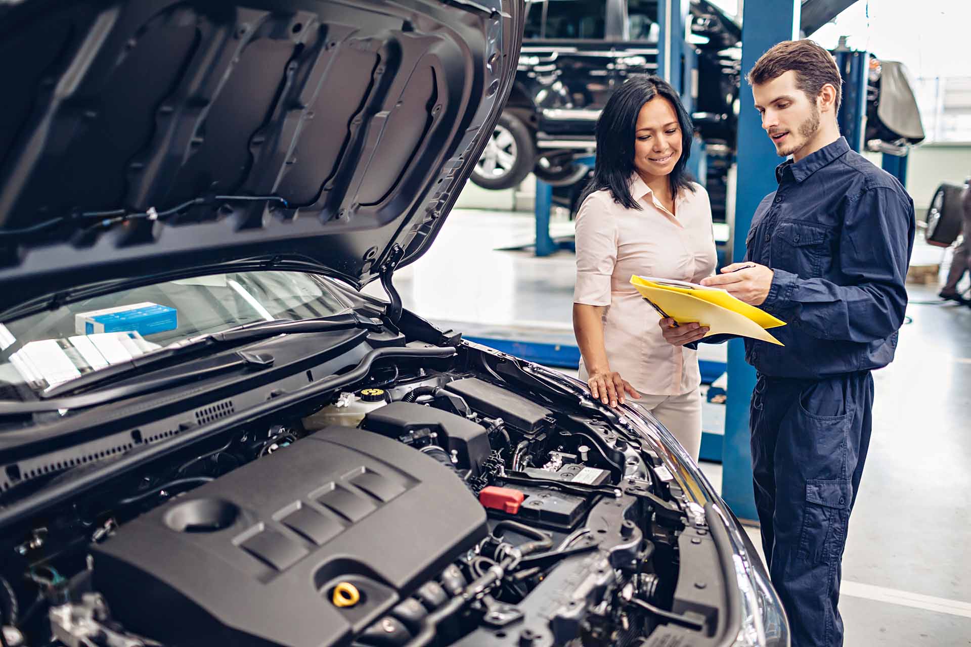 Mechanic talking to vehicle owner as they stand near car with hood up in body shop