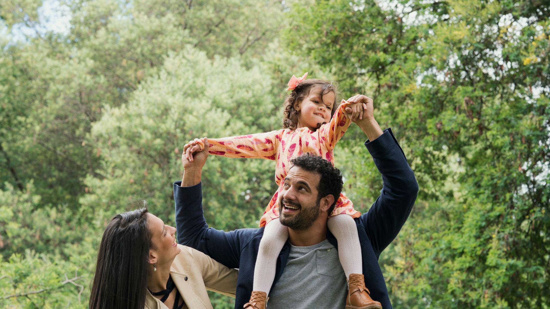 young girl sitting on fathers shoulders while mother smiles beside them