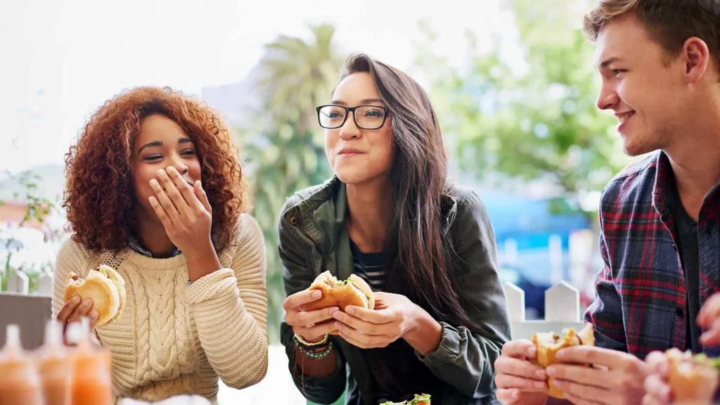 Three young adults laughing together while eating burgers outdoors