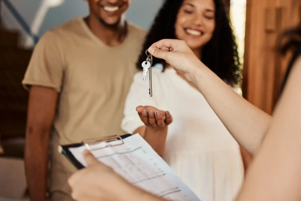 Hand passing house keys to a new homeowner with a smiling couple in the background