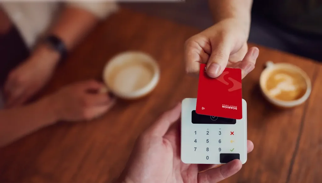 Hand tapping a red credit card on a contactless card reader at a cafe table