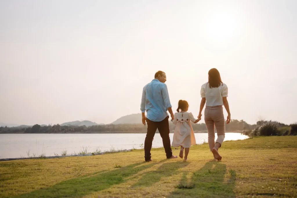 Family of three walking hand in hand toward a lake at sunset