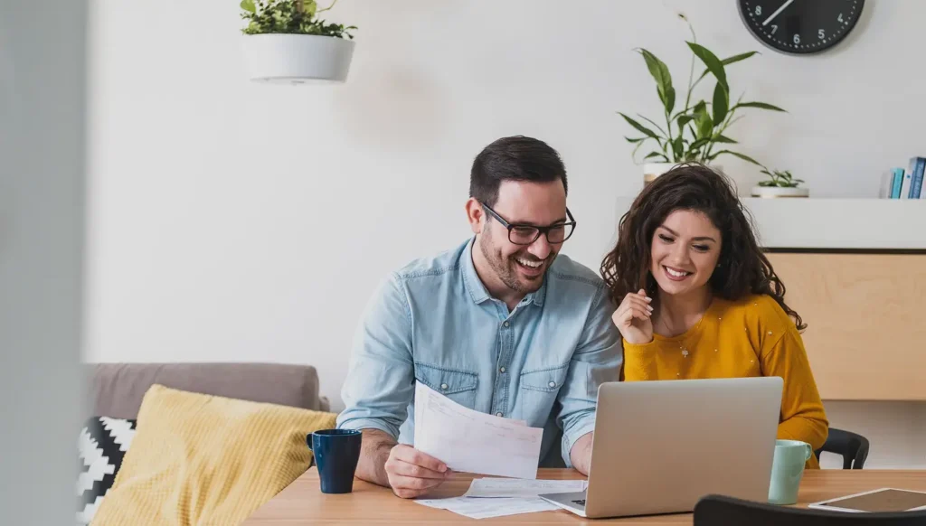 Couple smiling while reviewing paperwork together at a table with a laptop