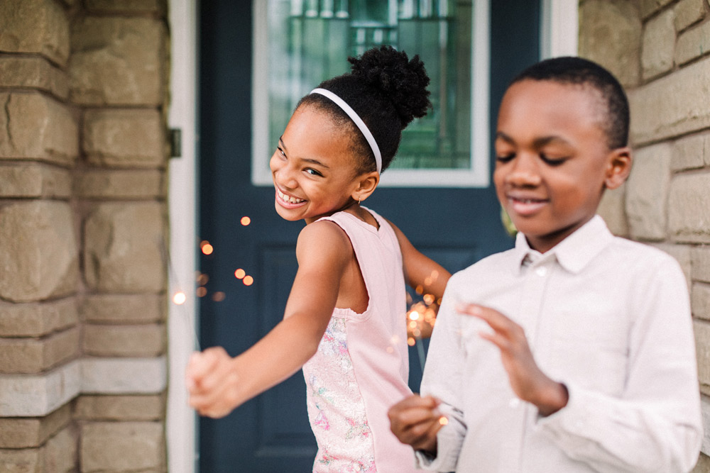 Two children smiling with sparklers outside the front door of a home